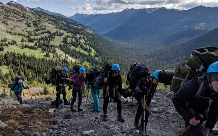A group of people wearing backpacks and helmets make their way along a rocky trail. Behind them is a vast mountainous valley.